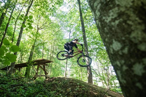A mountain biker catches air off a wooden jump on a forest trail, high above the ground.